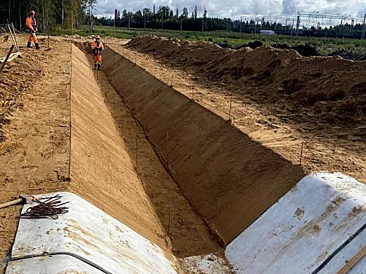 A trapezoidal drainage channel graded into sandy ground, setting-out pins along both crests and the first sheets laid at the near end