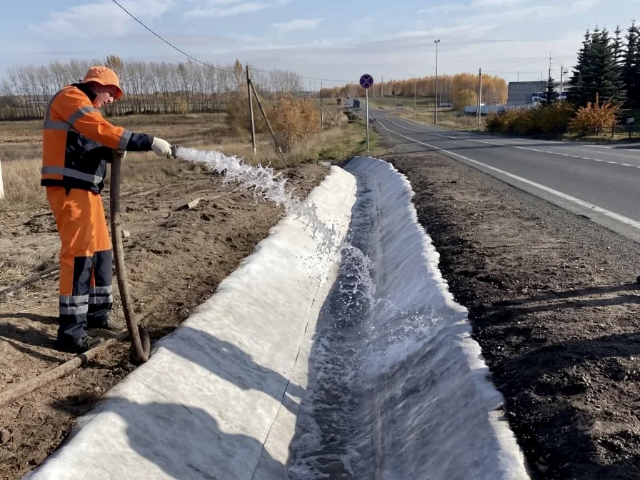 A worker spraying water along a roadside ditch lined with the material during hydration